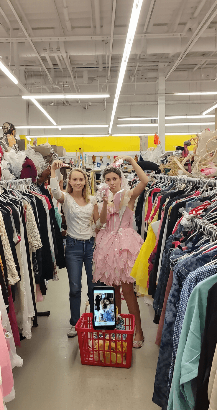 Family Halloween Costumes: Your Ultimate Guide to Creating Magic Together A brightly lit thrift store interior featuring a mother and daughter browsing costume racks, holding up various clothing items. Shopping baskets filled with budget-friendly pieces are nearby, and a smartphone shows reference photos of costume designs. Industrial-style lighting enhances the space, emphasizing the creative potential of second-hand finds.