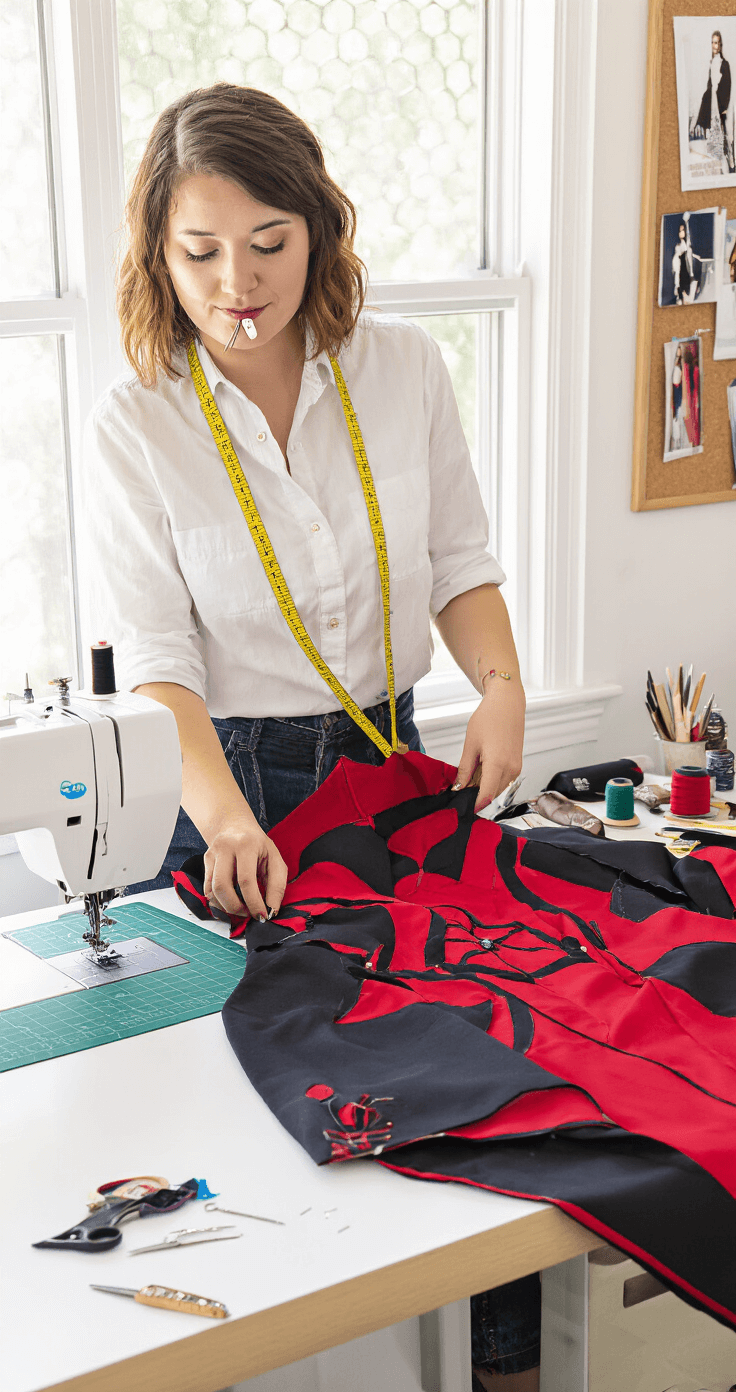 A female cosplayer works on custom modifications in a bright, organized sewing room, surrounded by sewing tools and an elaborate red and black costume in progress, illuminated by natural light.