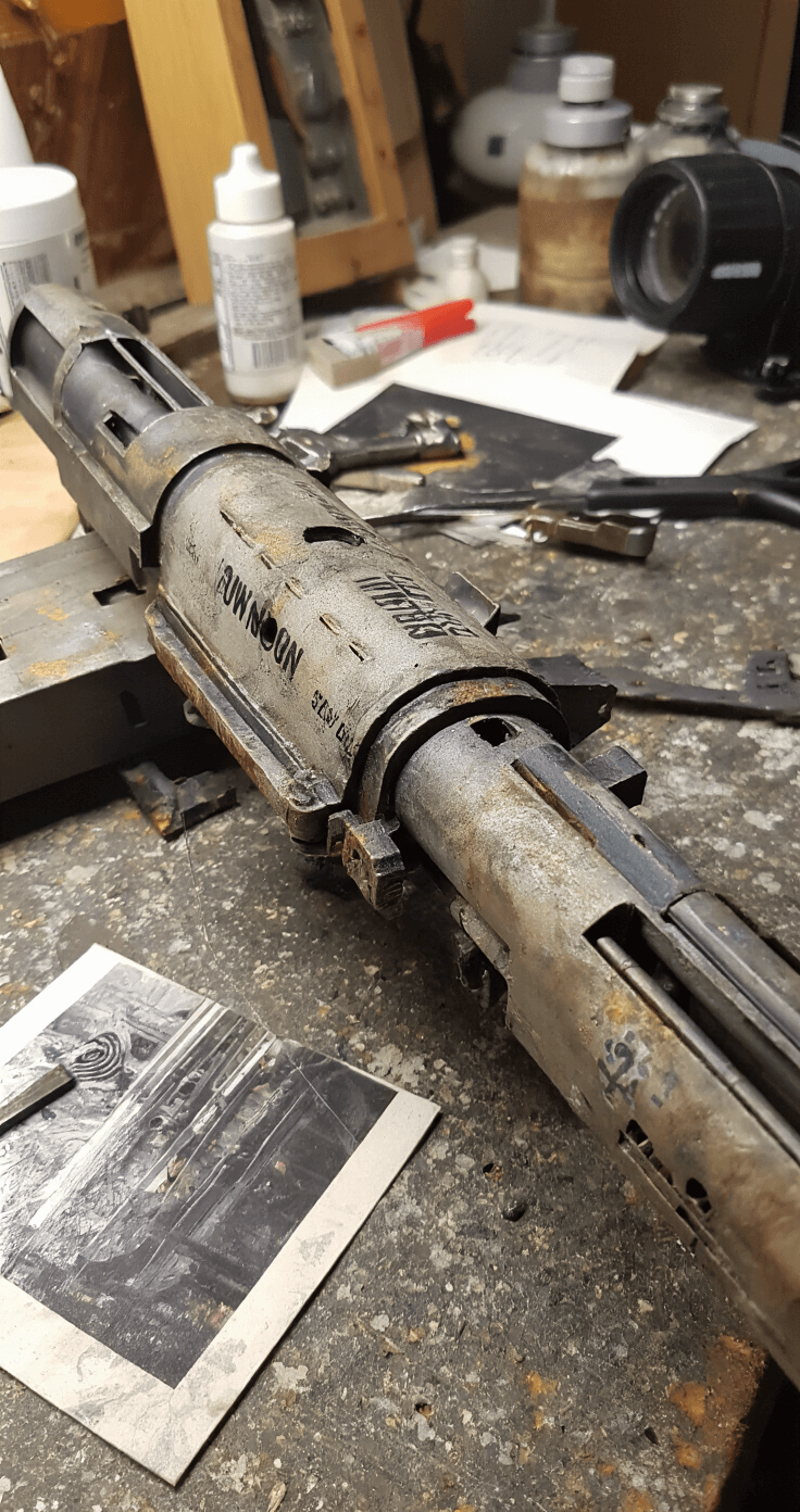 Creating the Perfect Jinx Cosplay: A Complete Guide for League of Legends Fans Close-up of a weathered Pow-Pow minigun prop on a workbench, showcasing metal effects, battle damage, and paint distressing illuminated by afternoon light. Various weathering tools and reference images are visible in the background, emphasizing detailed texture work.