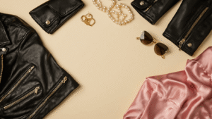 A flat lay of couples costume pieces featuring black leather jackets, pink satin accessories, vintage pearl jewelry, and retro sunglasses on a cream background, illuminated by soft golden hour lighting.