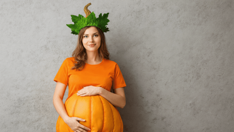 Pregnant woman wearing an orange maternity shirt styled as a pumpkin costume, adorned with a green leaf crown, on a clean background.