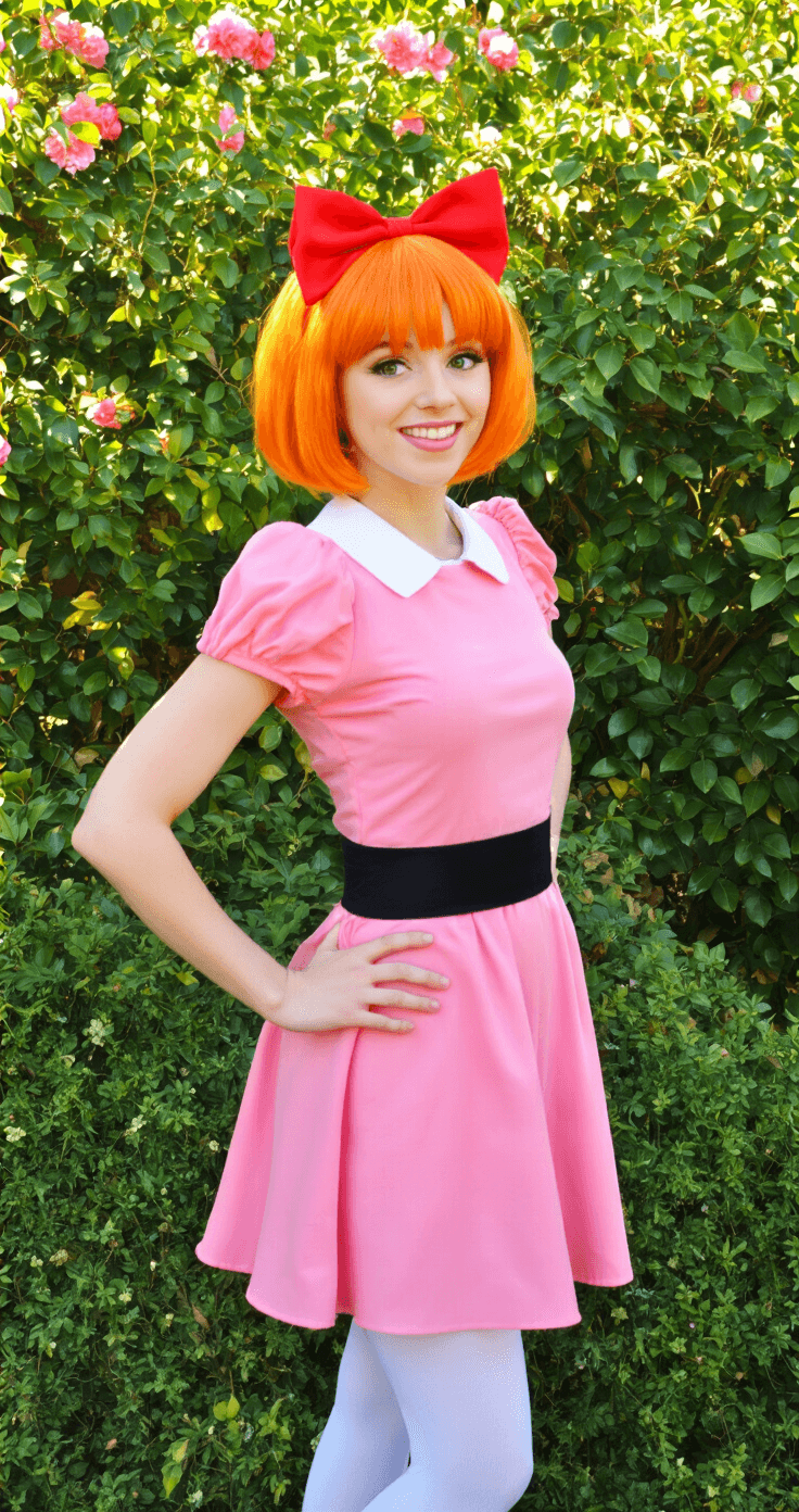 Orange Hair Cosplay: Your Ultimate Guide to Rocking Vibrant Character Looks A model in Powerpuff Girls Blossom cosplay poses in a sunlit garden, wearing a pink dress with a black belt, white tights, and an orange bob wig topped with a red bow, surrounded by flowering bushes and green foliage, with natural sunlight highlighting her vibrant hair.