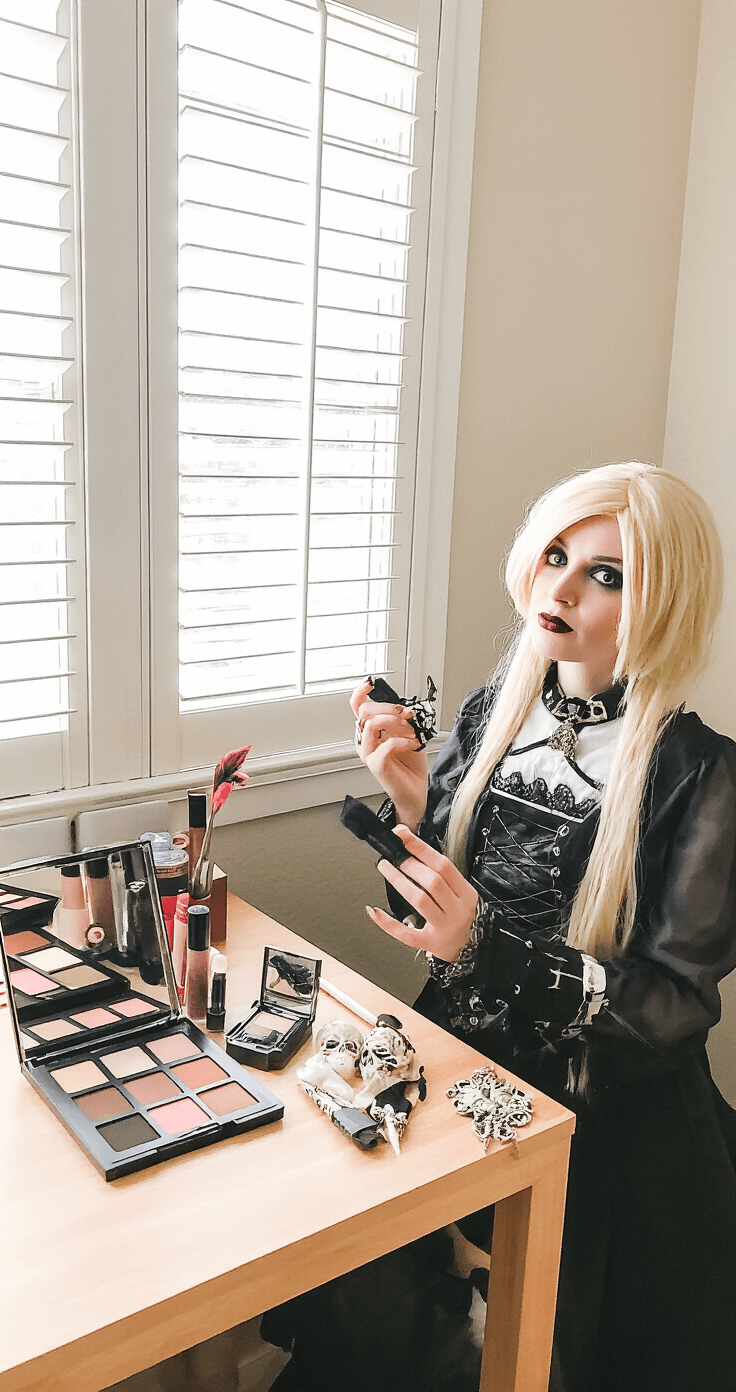 A budget-friendly bedroom setup with soft natural light, featuring affordable cosplay items like drugstore makeup, an inexpensive blonde wig, and DIY accessories on a wooden desk. A model showcases a homemade gothic outfit with handcrafted accessories, illustrating creativity and resourcefulness in cosplay.