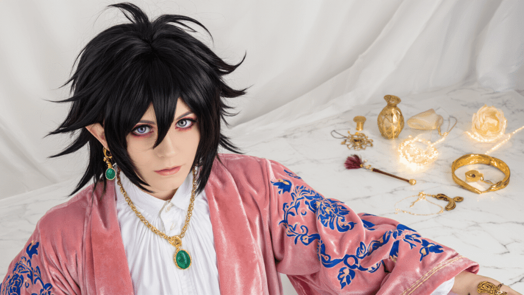 Dramatic black-haired cosplay wig and flowing pink coat with blue gold embroidery arranged on white marble, accompanied by a white poet shirt, emerald pendant, gold rings, and decorative ear clips, all bathed in warm golden hour lighting for an enchanting fantasy atmosphere.