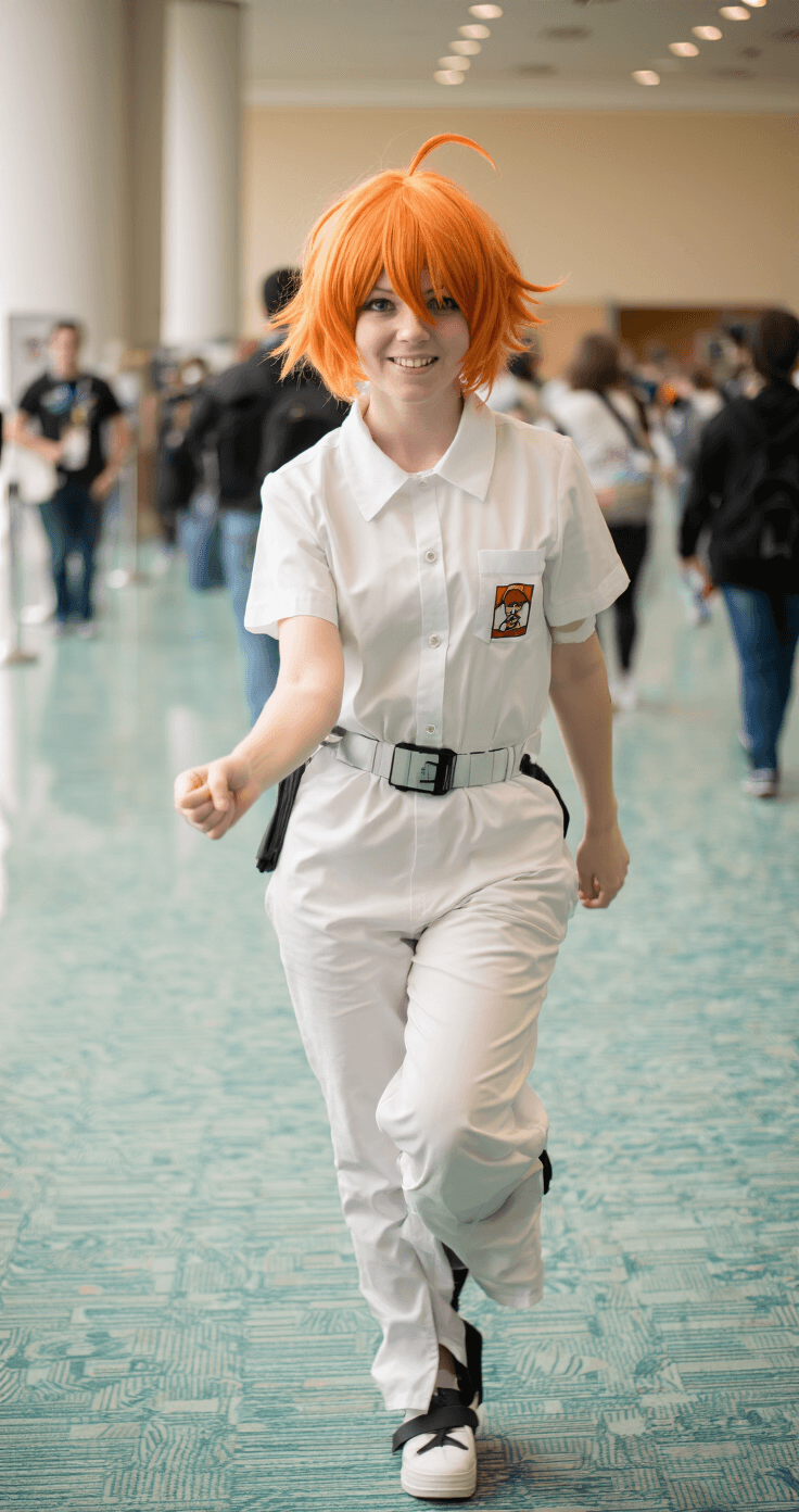 A dynamic cosplayer portraying Emma from The Promised Neverland, with short orange-blonde hair, wearing a prison uniform, captured mid-movement on a convention floor bathed in soft natural light, with a blurred background suggesting a lively atmosphere.