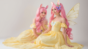 Fluttershy cosplay featuring a pastel yellow sundress, long pink wig, butterfly accessories, and cream-colored wings on a clean white background, capturing a whimsical and creative atmosphere.