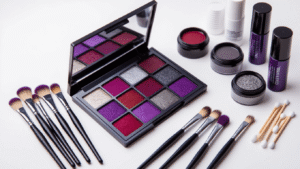 A well-organized makeup artist workspace displaying an open purple and black eyeshadow palette, neatly arranged makeup brushes, dark red and gray face paint tubes, metallic silver eyeshadow, black eyeliner pots, and scattered cotton swabs on a clean white surface, illuminated by soft, warm studio lighting.