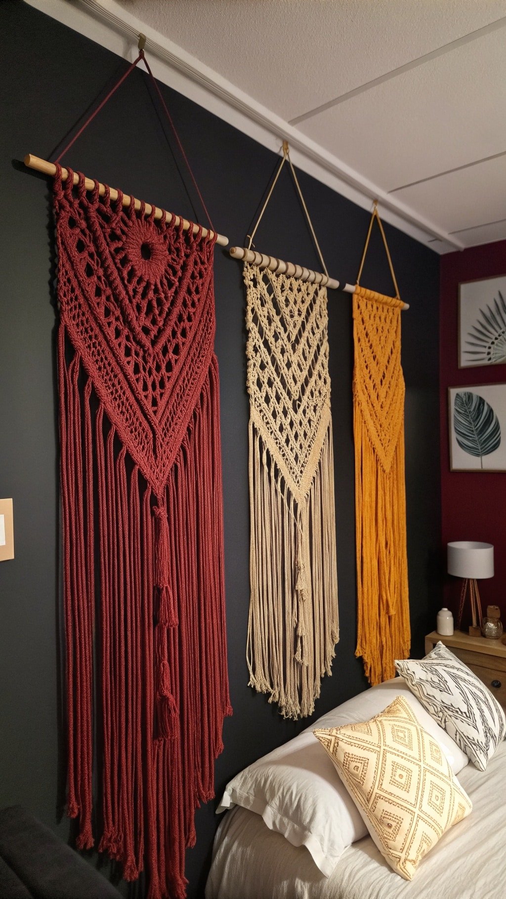 Three macramé wall hangings in red, beige, and orange against a dark wall in a boho bedroom.